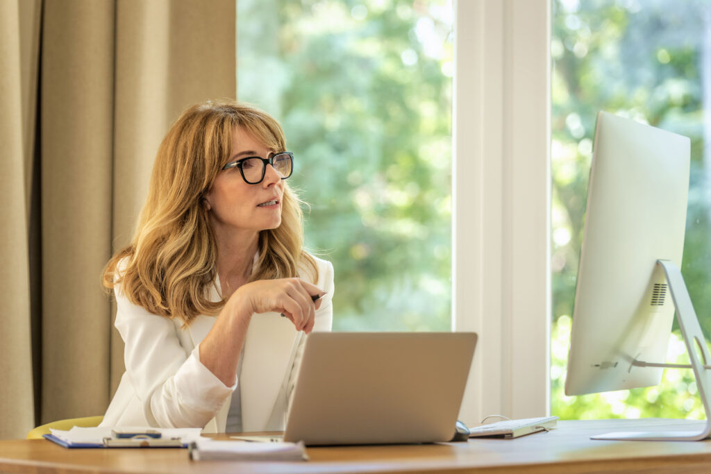 Cheerful smiling mid aged businesswoman sitting at desk and using laptop and computer for work. Confident professional female wearing white blazer and glasses.