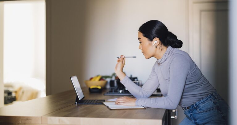 Beautiful Asian woman using digital tablet for online meeting while sitting in a modern kitchen.