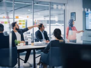 Multi-Ethnic Office Conference Room. Muslim Female CEO Wearing Hijab does Presentation for Group of Managers Talk, Use TV Infographics, Statistics. Businesspeople Brainstorm eCommerce Growth Strategy