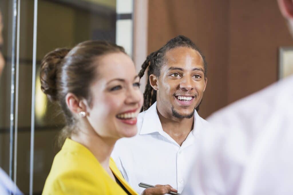 Close up of a multi-ethnic group of young business people in a meeting in an office. The focus is on a mixed race African American man with dreadlocks, wearing a white button-down shirt, smiling and looking at a businessman cropped and out of focus in the foreground.